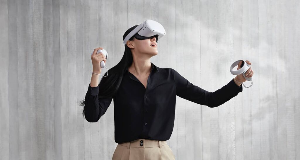 Woman plays Oculus Quest 2. She has an eyebox on and is holding hand sets in both hands. The design is sleek and white.