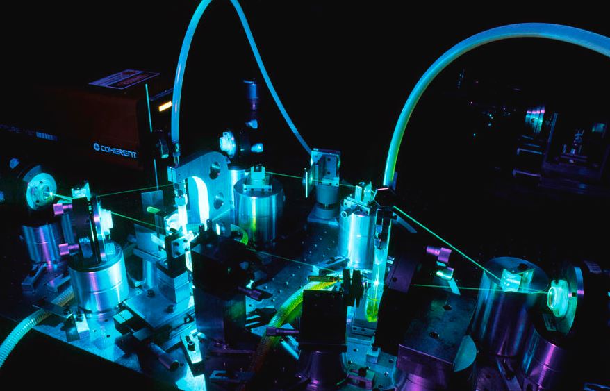 A dye laser in operation, with visible internal components including mounts, dye-carrying tubes, and laser beams, in a dimly lit room.