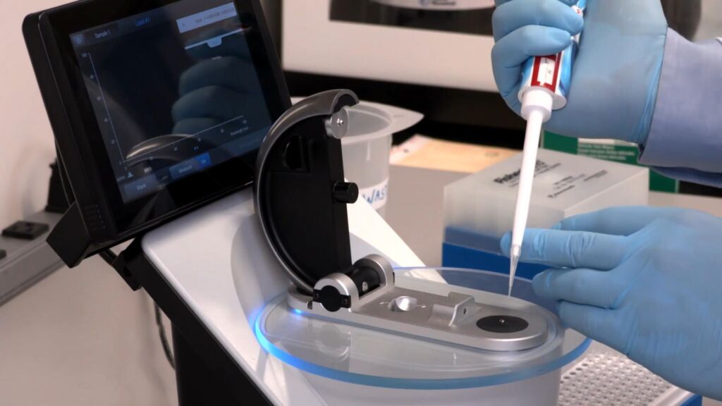 Microvolume Spectrophotometry: Lab technician's hand using a calibrated pipette to deposit a tiny droplet into the sample plate of a spectrophotometer system, demonstrating precise sample handling in microvolume spectrophotometry.